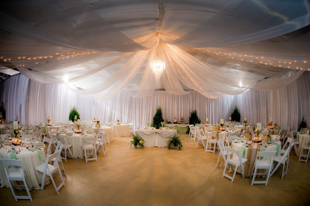 round white tables and chairs set up for wedding reception in room draped with white curtains and lights